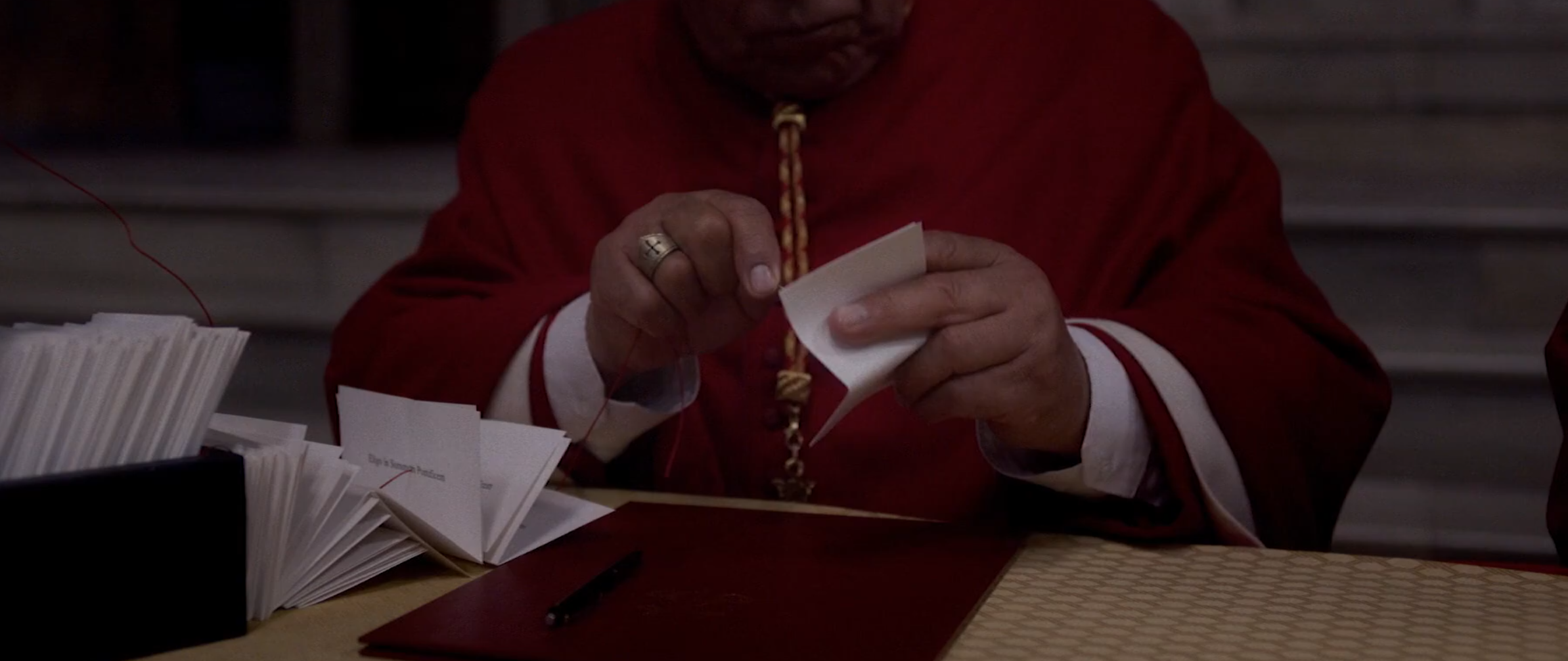 A still from the movie Conclave, in which a cardinal is piercing a folded piece of paper with a vote for pope on it with a needle, threading all the votes onto a red thread.